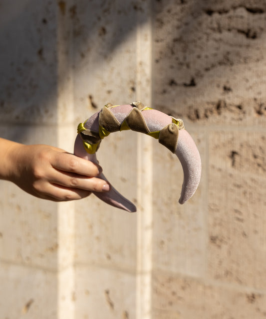 A hand displays the SAMPLE SALE Headband Knot Rose by WALD Berlin, a crescent-shaped pink accessory with green accents and triangular details, set against a textured beige wall.