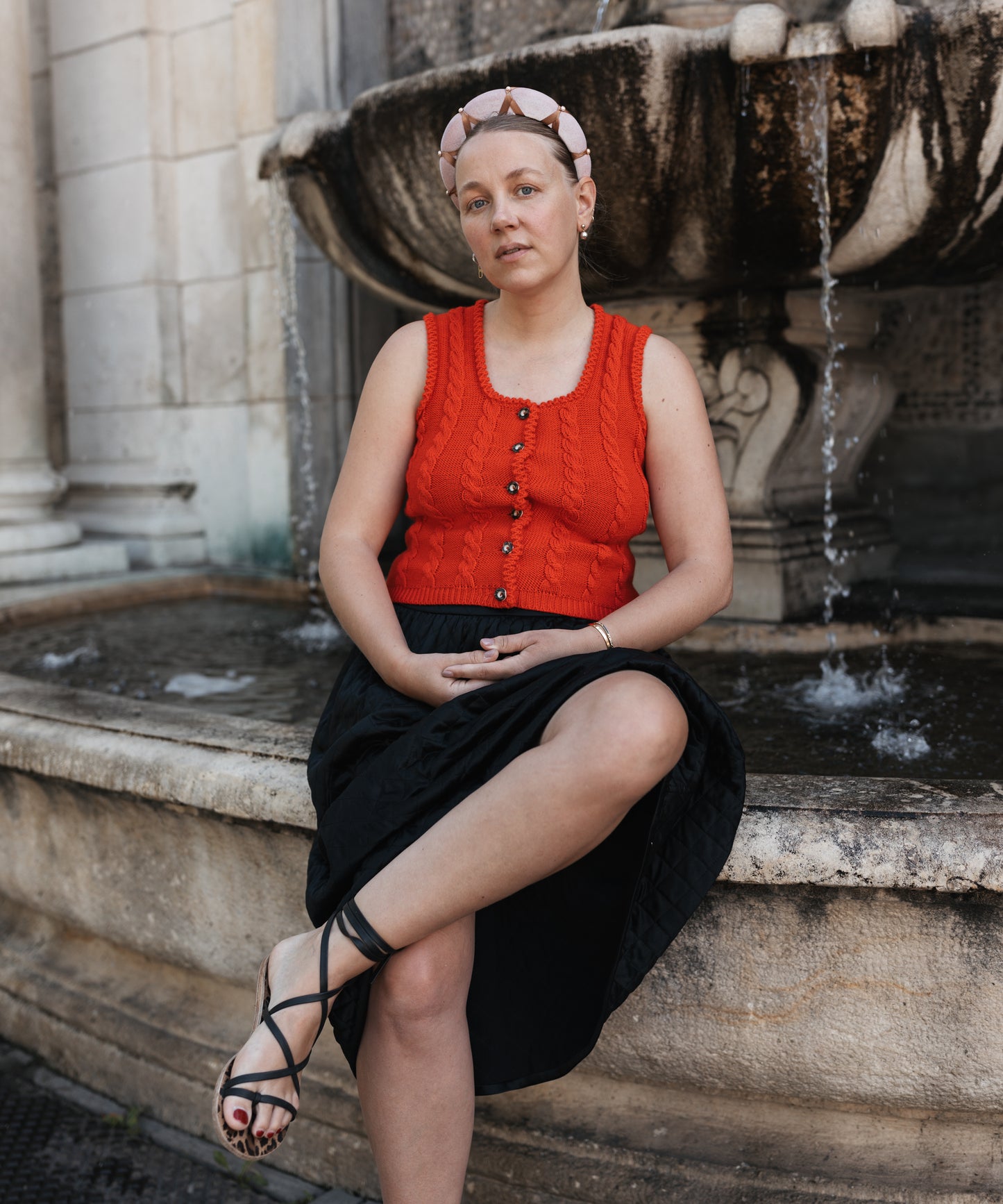 Wearing the WALD Berlin SAMPLE SALE Headband Knot Rose, a person in a red sleeveless top, black skirt, and sandals sits by a stone fountain with water flowing in the background.