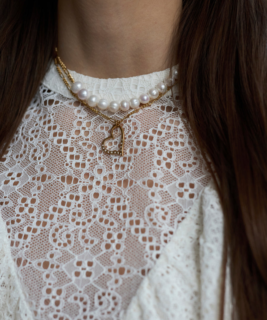 A person wearing a white lace top and two necklaces: a pearl necklace and the WALD Berlin SAMPLE SALE Valentine’s Day special sketch heart necklace, plated in 18k gold and adorned with sparkling Swarovski stones.