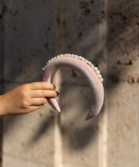 A hand displays the SAMPLES SALE Lady Di Headband Rose by WALD Berlin—a crescent-shaped, light pink velvet headband embellished with pearls—handmade in Germany against a sunlit, textured wall.