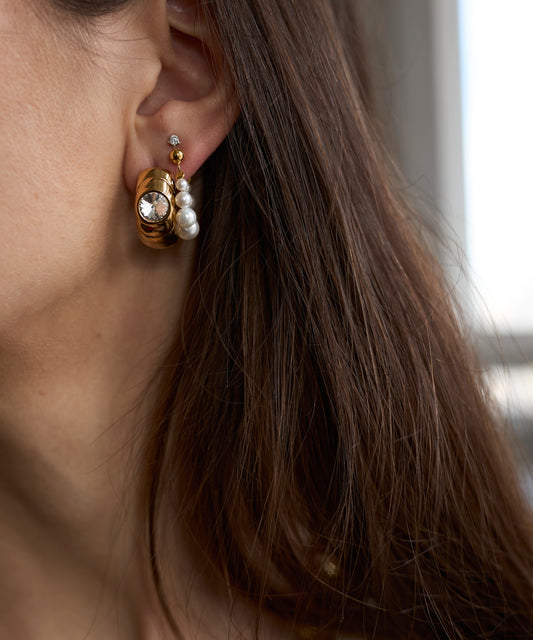 Close-up of a woman's ear with three earrings, including the Isla bonita ready or not earring by WALD Berlin. The design features gold plating and white pearls, while brown hair partially covers the ear.