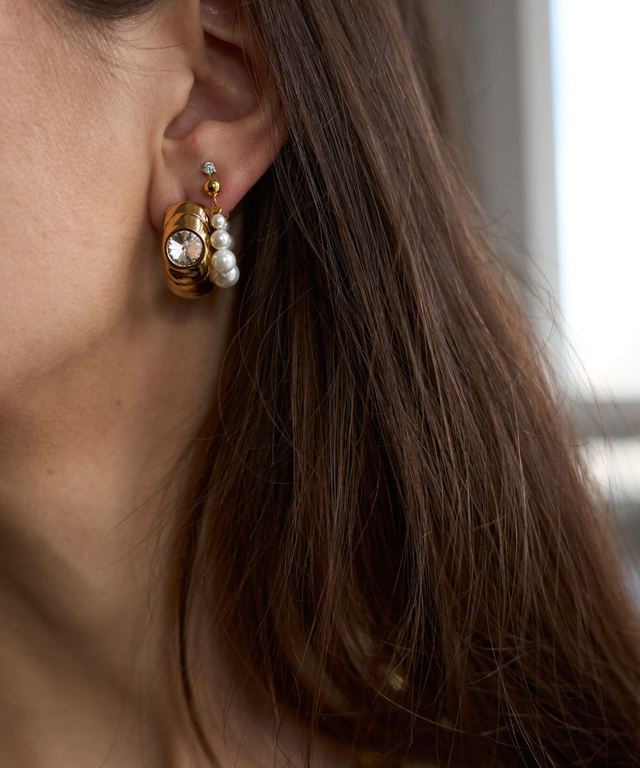 Close-up of a woman's ear with three earrings, including the Isla bonita ready or not earring by WALD Berlin. The design features gold plating and white pearls, while brown hair partially covers the ear.