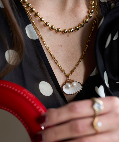 Close-up of a person wearing the WALD Berlin Harry Necklace Gold, layered with other gold-plated jewelry, including a shell and pearl piece and a gold ring, paired with a black polka dot top. Some pieces are made in Germany.