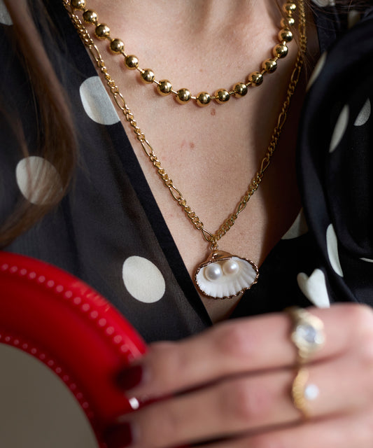 Close-up of a person wearing the WALD Berlin Harry Necklace Gold, layered with other gold-plated jewelry, including a shell and pearl piece and a gold ring, paired with a black polka dot top. Some pieces are made in Germany.