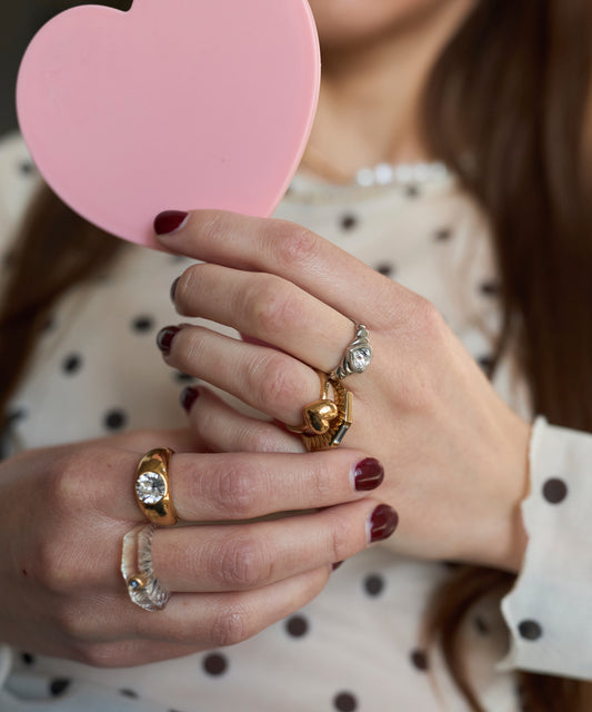 A person with maroon nail polish holds a pink heart-shaped mirror, showcasing elegant ring stacking featuring the WALD Berlin Shining Star Gold Ring on their fingers.