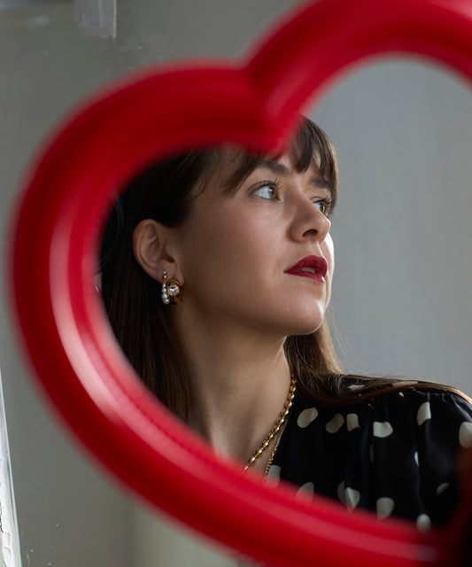 A woman wearing red lipstick and WALD Berlin's Isla bonita ready or not earrings is seen through a red heart-shaped frame, glancing sideways against a neutral backdrop.
