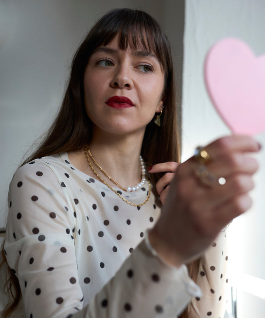 A woman in a white polka dot blouse holds a pink heart-shaped mirror, wearing WALD Berlin’s Exclusive Heart Black Earrings and red lipstick; her gold jewelry adds elegance as she admires her reflection.