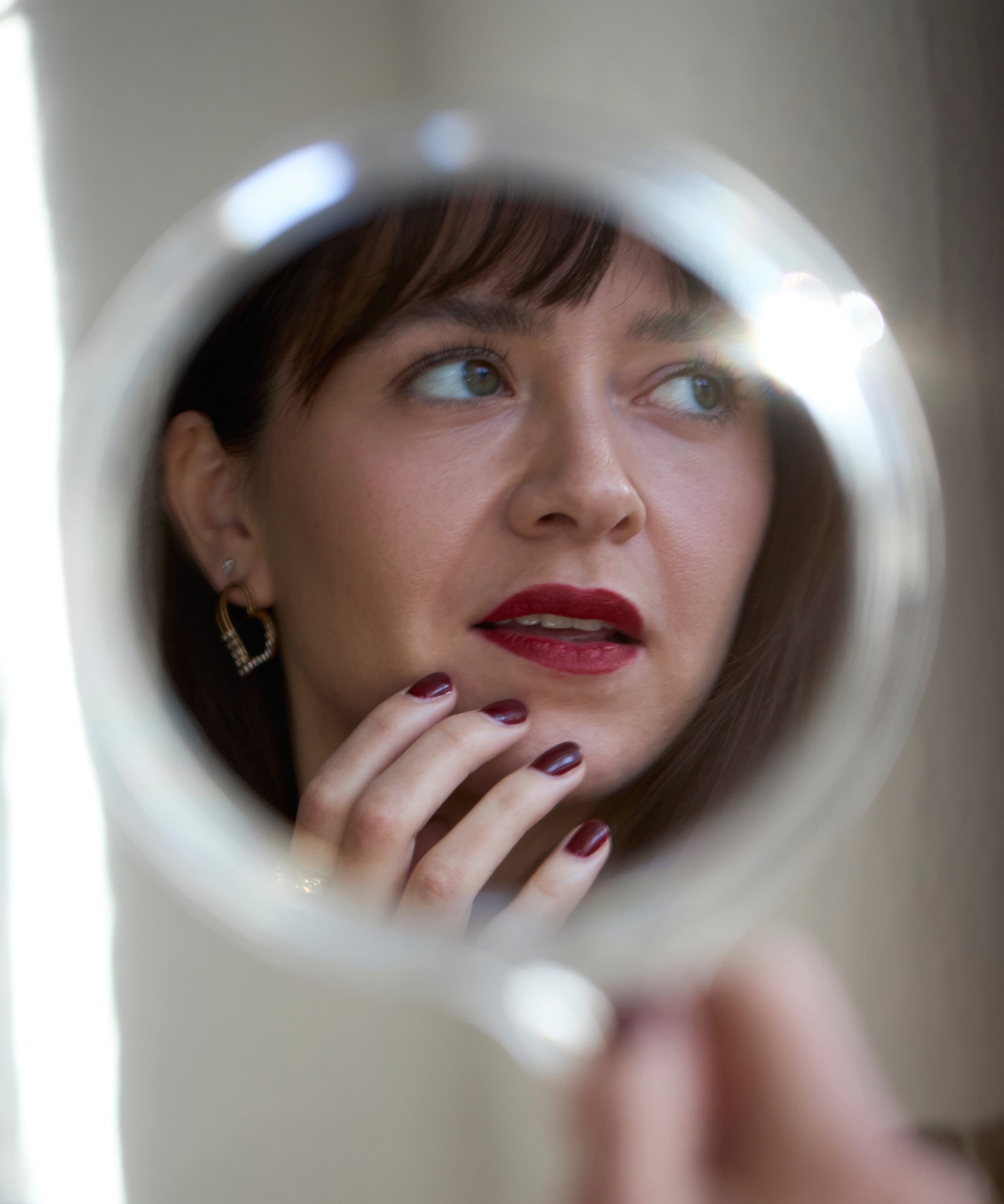 A woman with red lipstick and dark nail polish touches her chin, her reflection showing WALD Berlin Valentines Day special sketch heart earrings sparkling with Swarovski stones.