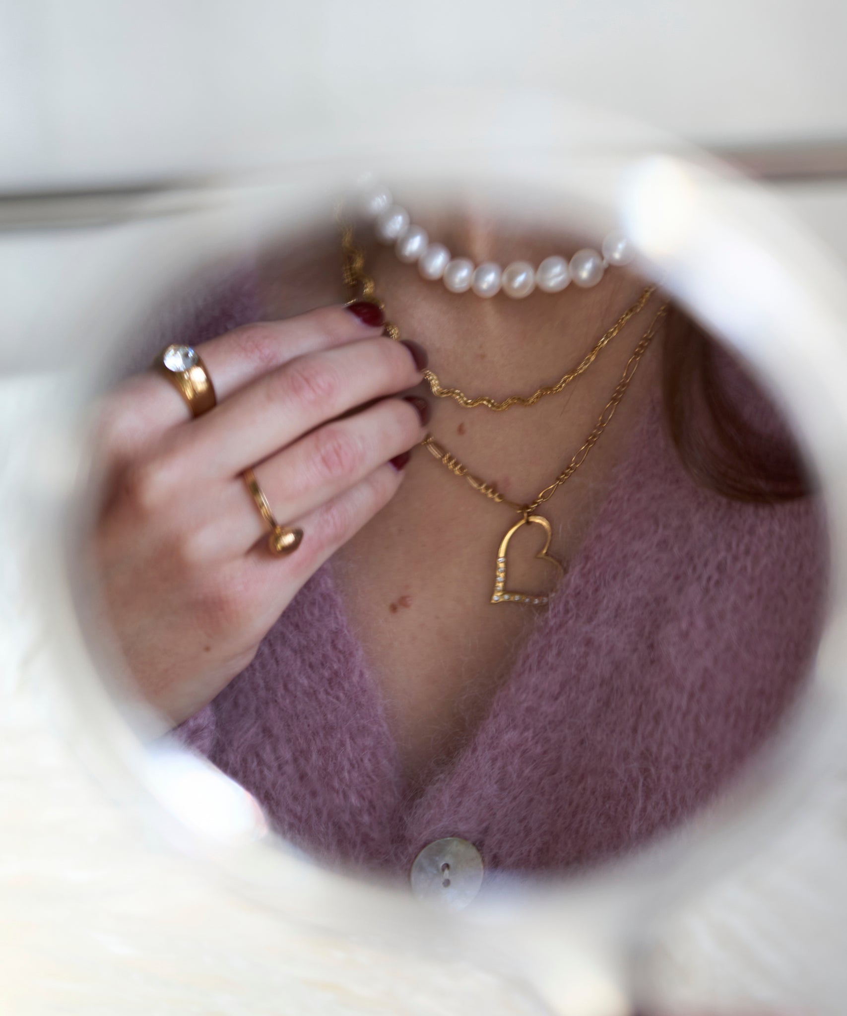 A close-up shows a person wearing layered necklaces, including the WALD Berlin Valentine’s Day special sketch heart necklace, with rings on their fingers, all reflected in a round mirror.