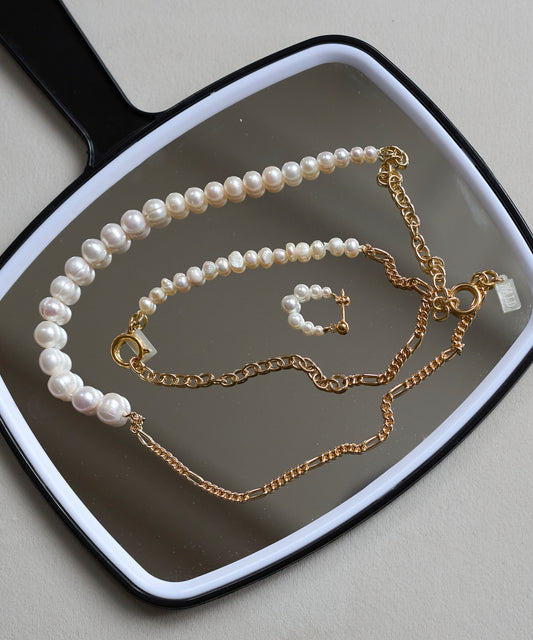 A handheld mirror displays WALD Berlin’s Isla Bonita Pearl Hoops, a pearl necklace, a pearl bracelet with a gold cross charm, and two gold plated sterling silver chain necklaces.