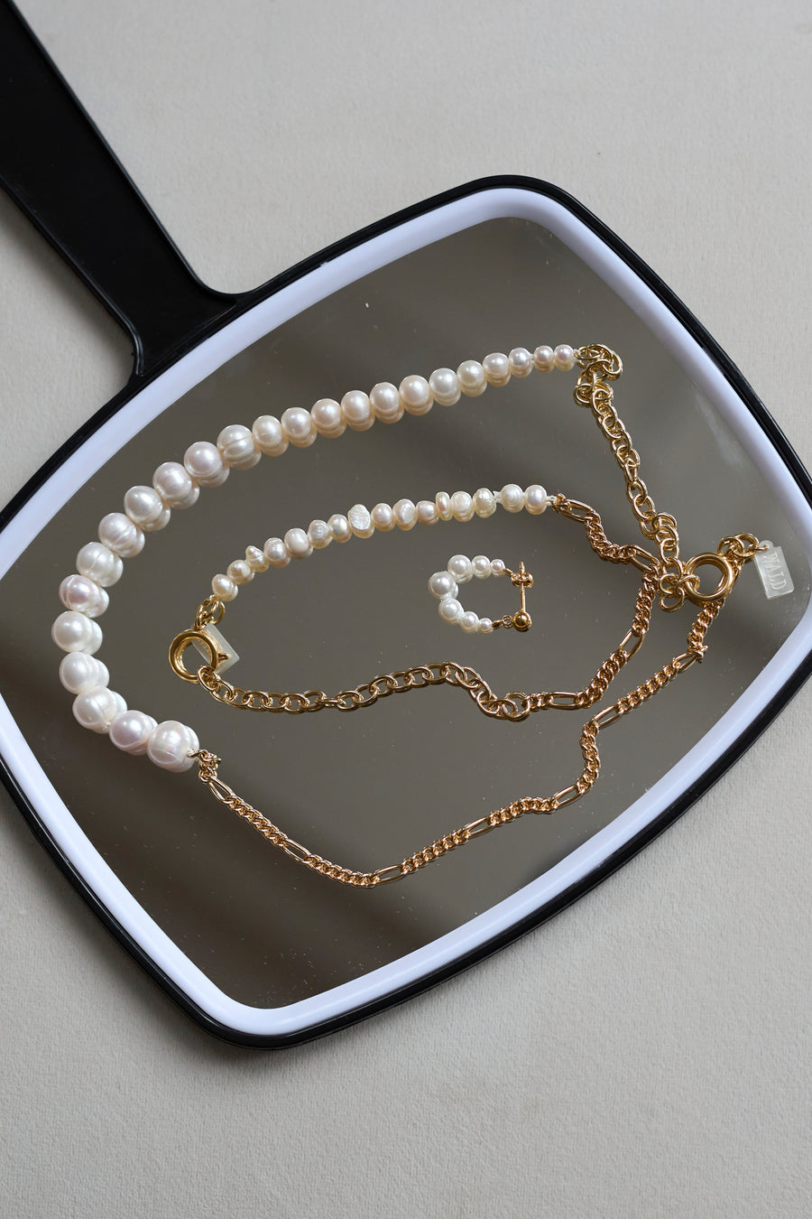 A handheld mirror displays WALD Berlin’s Isla Bonita Pearl Hoops, a pearl necklace, a pearl bracelet with a gold cross charm, and two gold plated sterling silver chain necklaces.