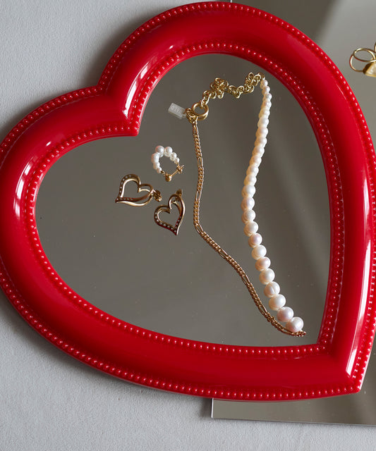 A red heart-shaped mirror displays a pearl necklace, an 18k gold-plated chain, and WALD Berlin’s Valentines Day special sketch heart earrings.