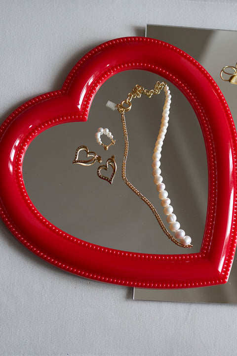 A red heart-shaped mirror displays a pearl necklace, an 18k gold-plated chain, and WALD Berlin’s Valentines Day special sketch heart earrings.
