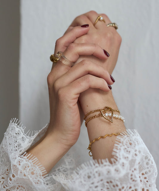 Two hands with dark red nail polish are clasped, displaying 18k gold-plated rings and the WALD Berlin Valentines Day special sketch heart bracelet adorned with Swarovski stones over white lace sleeves on a light background.
