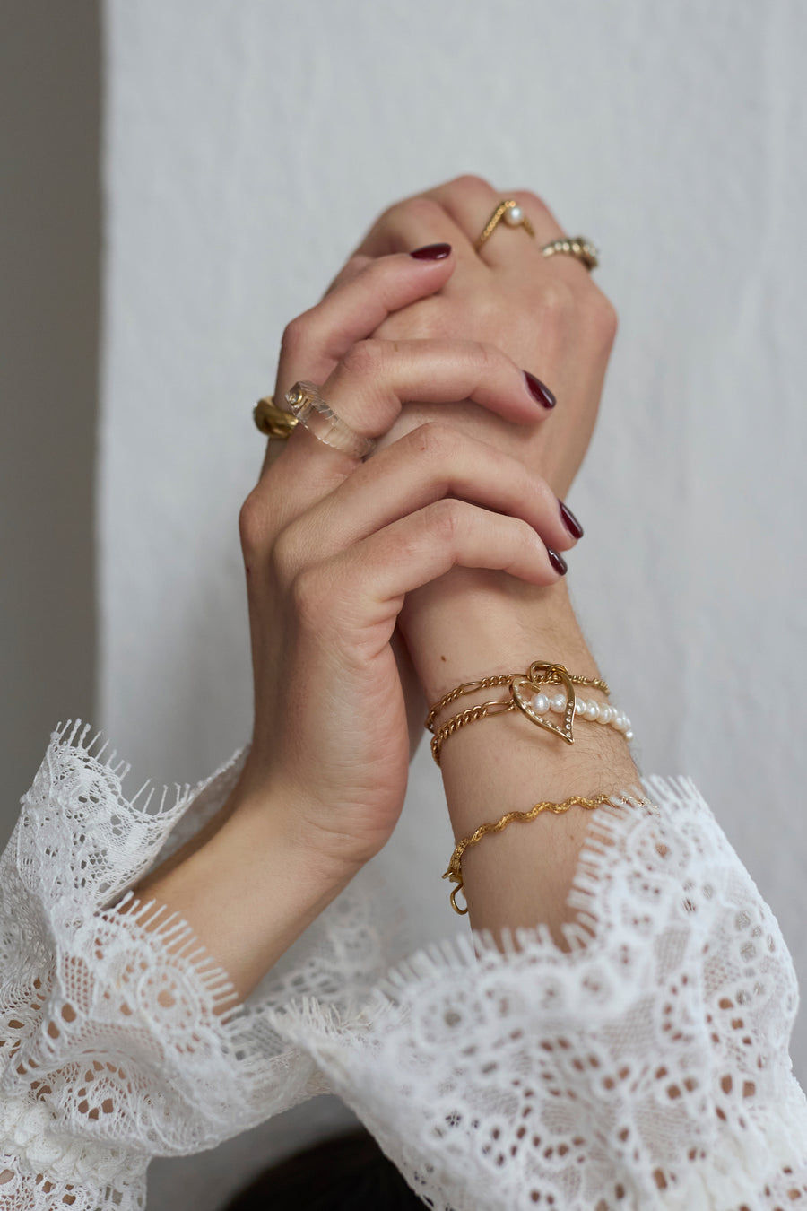 Two hands with burgundy nail polish are clasped, adorned with WALD Berlin gold rings and a SAMPE SALE Valentines Day special sketch heart bracelet; gold-plated bracelets and white lace sleeves enhance the elegant look.