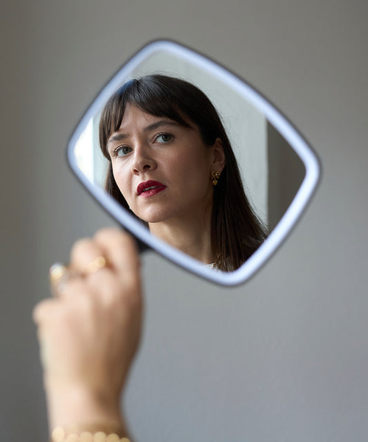 A woman with dark hair and red lipstick is reflected in a handheld mirror, holding up her hand adorned with 18k gold-plated jewelry and WALD Berlin’s Valentines Day special bow heart earrings sparkling with Swarovski stones.