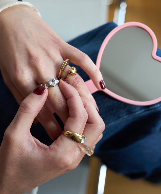 Close-up of manicured hands wearing WALD Berlin’s Mom Heart Ring, a gold-plated brass piece, beside a pink heart-shaped mirror on denim.