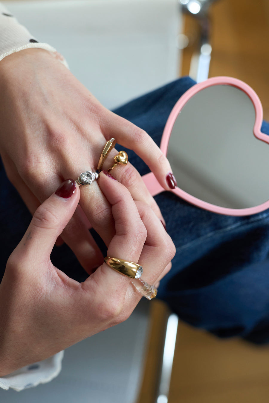 Close-up of manicured hands wearing WALD Berlin’s Mom Heart Ring, a gold-plated brass piece, beside a pink heart-shaped mirror on denim.