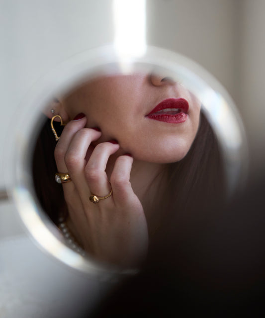 A woman's face with red lipstick and gold jewelry is reflected in a round mirror, her hand—adorned with the WALD Berlin Mom Heart Ring—gently touching her cheek.