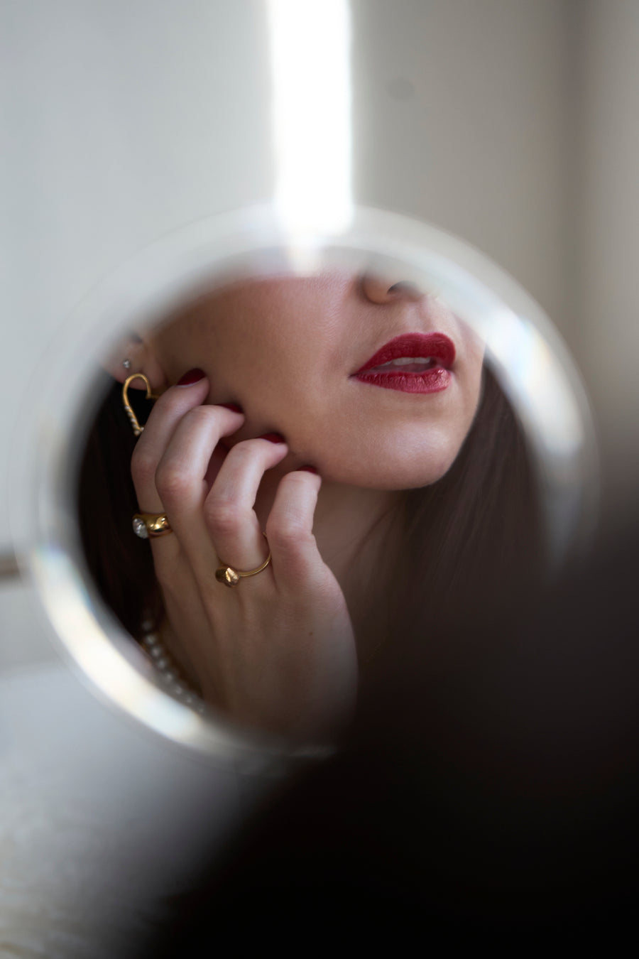 A woman's face with red lipstick and gold jewelry is reflected in a round mirror, her hand—adorned with the WALD Berlin Mom Heart Ring—gently touching her cheek.