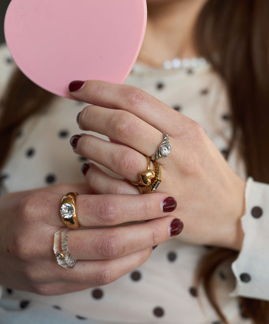 Close-up of a person with maroon nails holding a pink heart-shaped object, wearing multiple love-themed jewelry pieces including the WALD Berlin Mom Heart Ring, and dressed in a white polka dot blouse.