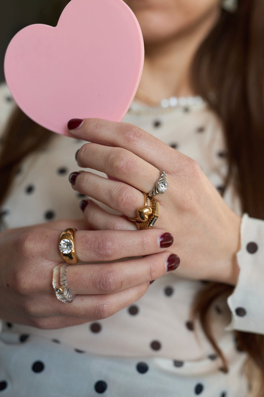 Close-up of a person with maroon nails holding a pink heart-shaped object, wearing multiple love-themed jewelry pieces including the WALD Berlin Mom Heart Ring, and dressed in a white polka dot blouse.