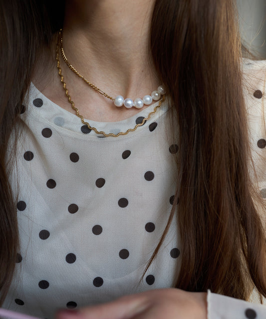 A woman with long brown hair wears a white blouse with black polka dots and layered necklaces, including the WALD Berlin Irina Necklace Gold.