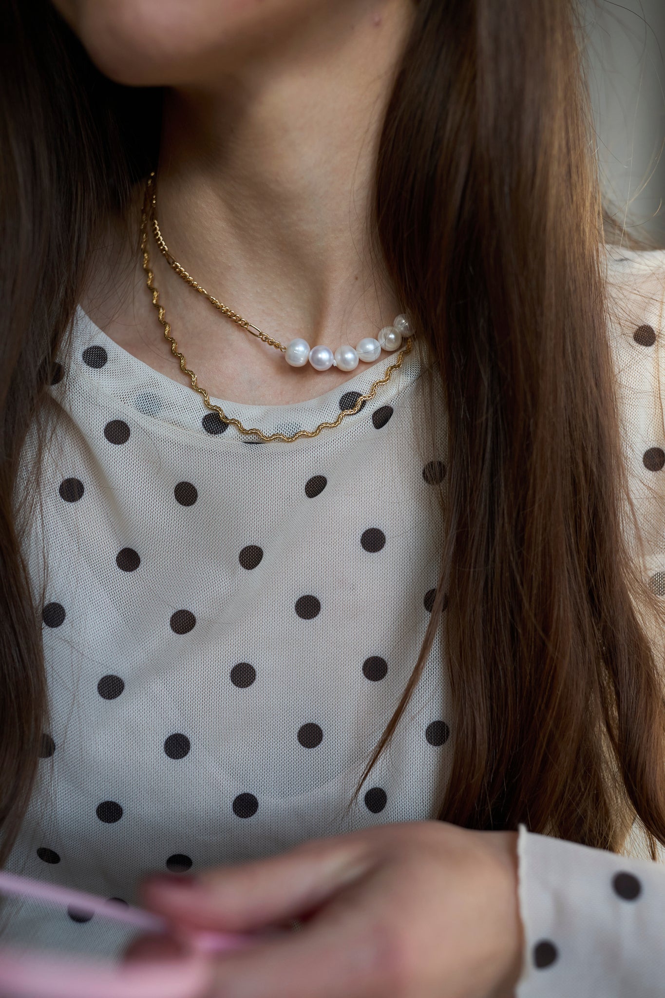 A woman with long brown hair wears a white blouse with black polka dots and layered necklaces, including the WALD Berlin Irina Necklace Gold.