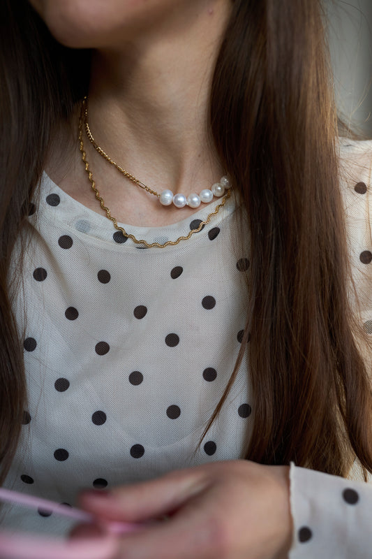 A woman with long brown hair wears a white blouse with black polka dots and layered necklaces, including the WALD Berlin Irina Necklace Gold.