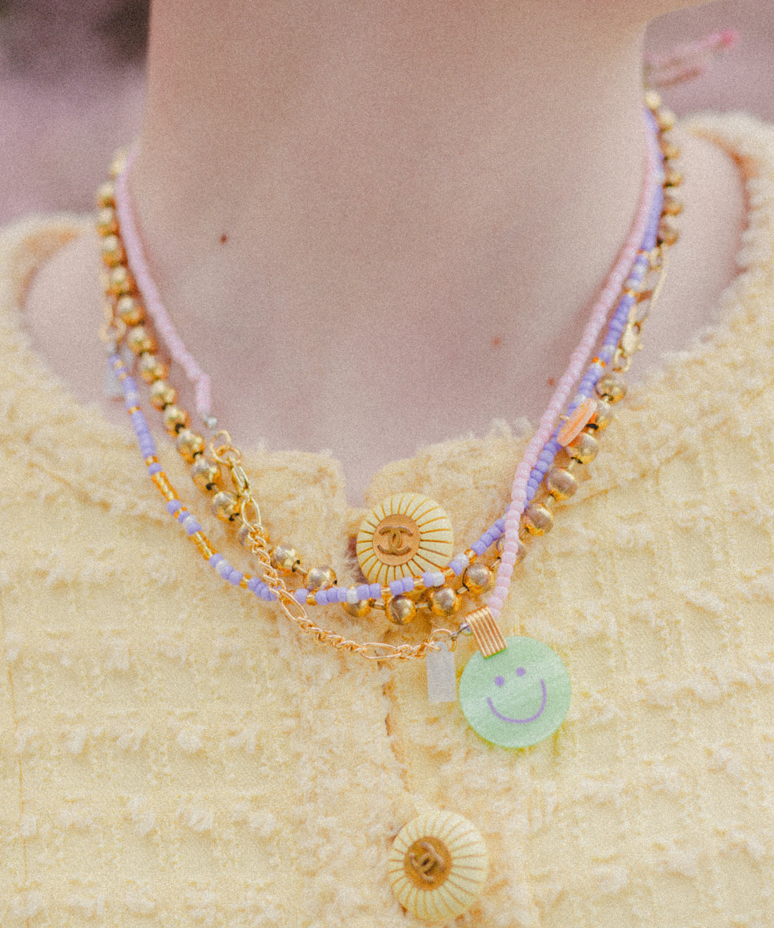 A person wears a yellow textured garment with layered necklaces, including beaded 18k gold plated pieces and WALD Berlin's Pop Smilie Mini necklace featuring a green Smilie Medaillon pendant.