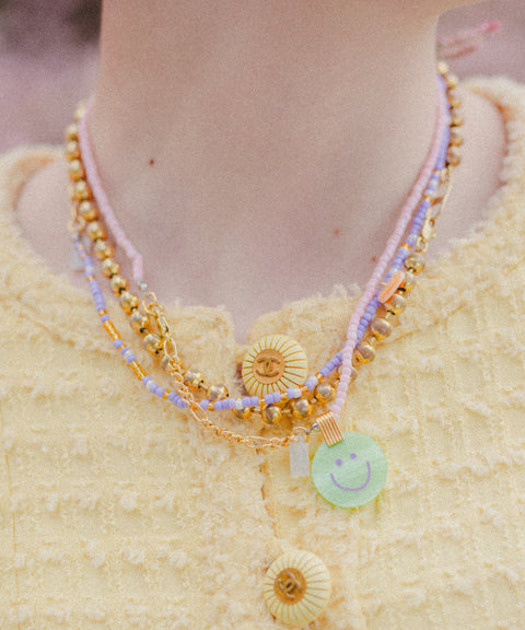 A person wears a yellow textured garment with layered necklaces, including beaded 18k gold plated pieces and WALD Berlin's Pop Smilie Mini necklace featuring a green Smilie Medaillon pendant.
