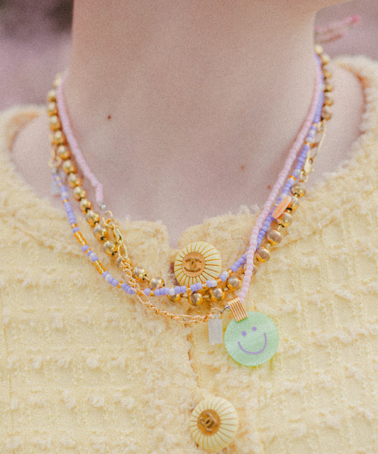 A person wears a yellow textured garment with layered necklaces, including beaded 18k gold plated pieces and WALD Berlin's Pop Smilie Mini necklace featuring a green Smilie Medaillon pendant.