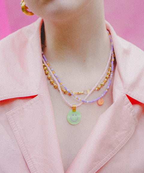 Person wearing a pale pink collared jacket and layered colorful necklaces, including the WALD Berlin Pop Smilie Mini 18k gold plated medallion, against a pink background.