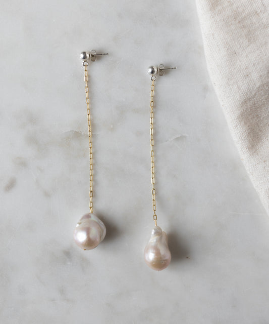 A pair of WALD Berlin's Happy Holidays Pearl Drop Earrings, featuring a gold chain design with large, irregularly shaped pearls, displayed on a light marble surface.