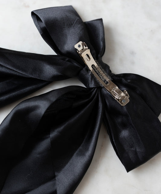 The Happy Holidays Black Hair Bow by WALD Berlin, a large satin hair clip ideal for festive styling, is showcased on a white marble surface.