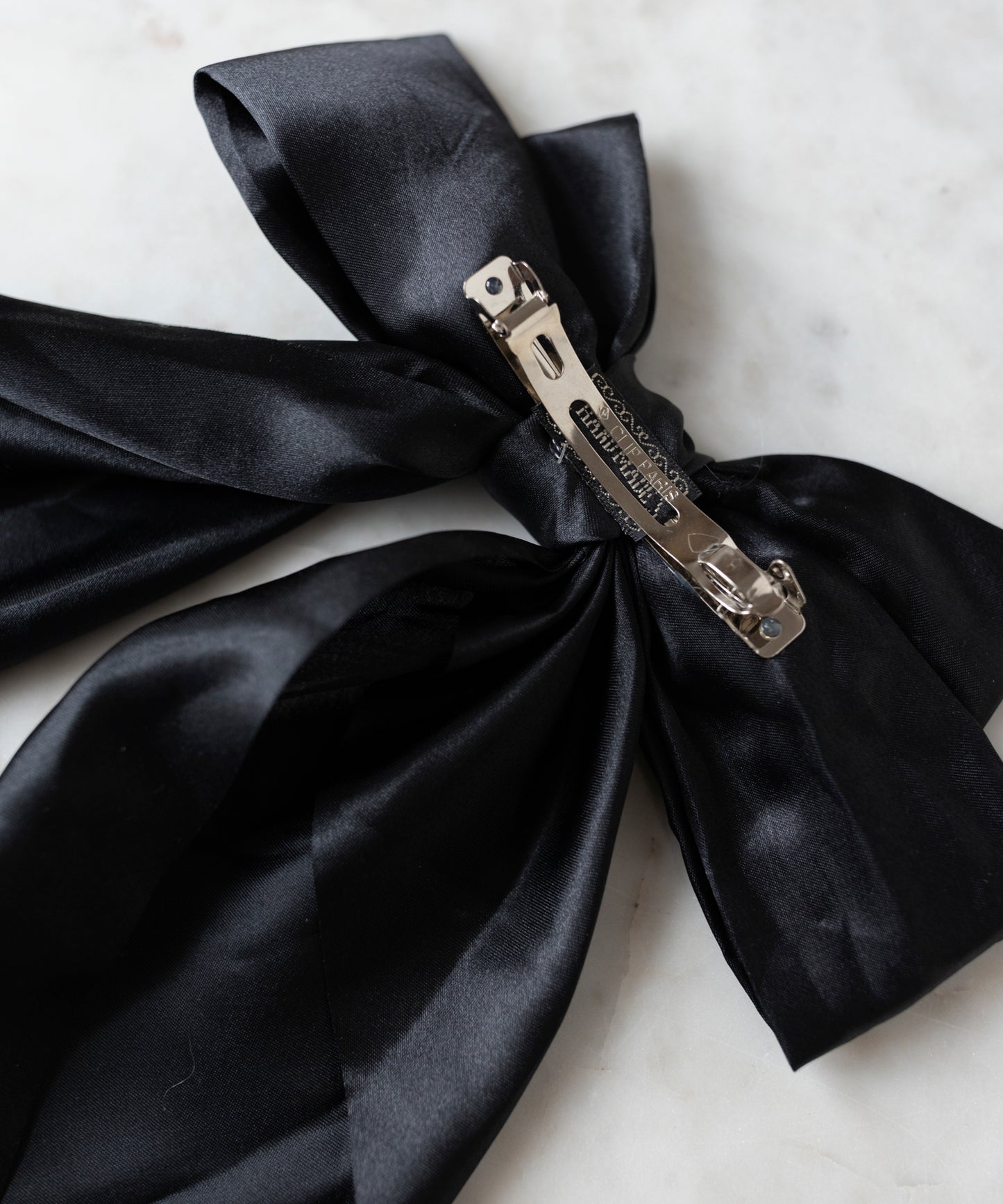 The Happy Holidays Black Hair Bow by WALD Berlin, a large satin hair clip ideal for festive styling, is showcased on a white marble surface.