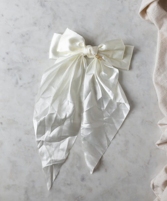The Happy Holidays Cream Hair Bow by WALD Berlin rests elegantly on a light marble surface, its long, flowing ends cascading downward, making it perfect for festive styling.
