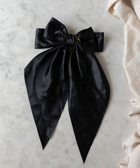 The Happy Holidays Black Hair Bow by WALD Berlin, featuring a large satin design with long tails and a gold pin, is gracefully displayed on a light marble surface—ideal for holiday hair or festive styling.