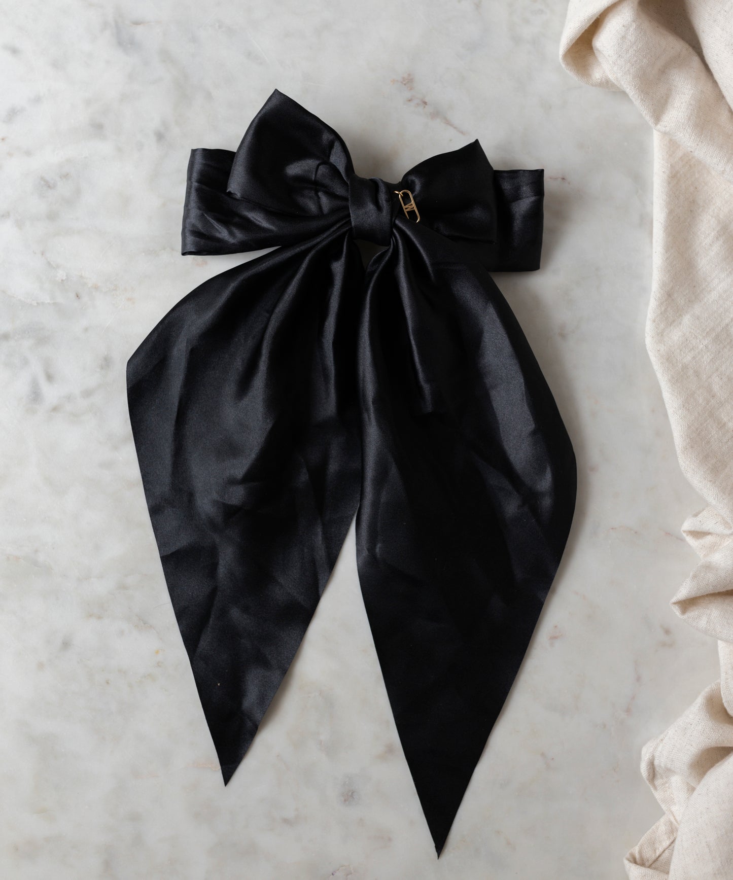 The Happy Holidays Black Hair Bow by WALD Berlin, featuring a large satin design with long tails and a gold pin, is gracefully displayed on a light marble surface—ideal for holiday hair or festive styling.