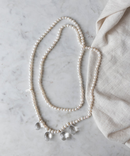 The Happy Holidays Long Pearl Drop Necklace by WALD Berlin, adorned with sweetwater pearls and three glass pendants, is gracefully displayed on a marble surface beside a piece of beige fabric.