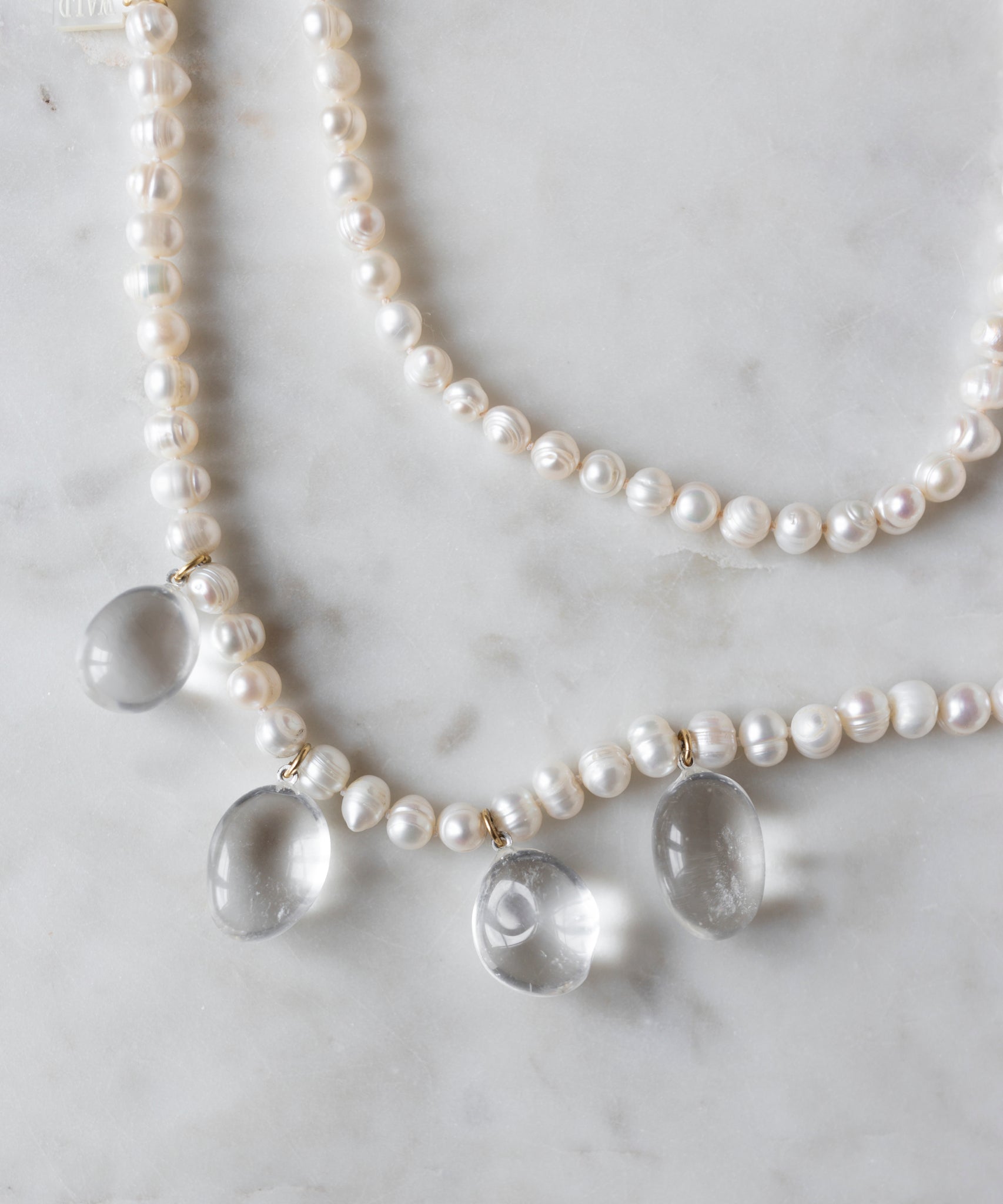 The Happy Holidays Long Pearl Drop Necklace by WALD Berlin gracefully lies on the light gray marble surface, adorned with sweetwater pearls and sparkling berg crystal pendants.