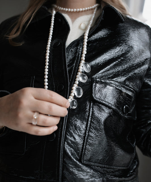 A person wearing a black leather jacket holds the Happy Holidays Long Pearl Drop Necklace from WALD Berlin, which is adorned with sweetwater pearls, clear beads, and a ring.