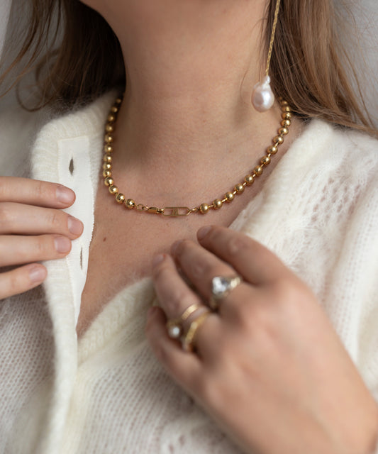 A person adorned with the Harry Necklace Gold by WALD Berlin, a gold beaded necklace, alongside multiple rings on their fingers and an 18k gold-plated brass bracelet, dressed in a white knitted sweater.