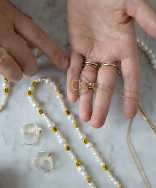 A hand is holding a WALD Berlin Smilie Dude Medaillon Gold Necklace, adorned with a gold crescent moon pendant. Various pieces of jewelry, including hand-painted polyester and pearl bead necklaces, are displayed on the marble surface.