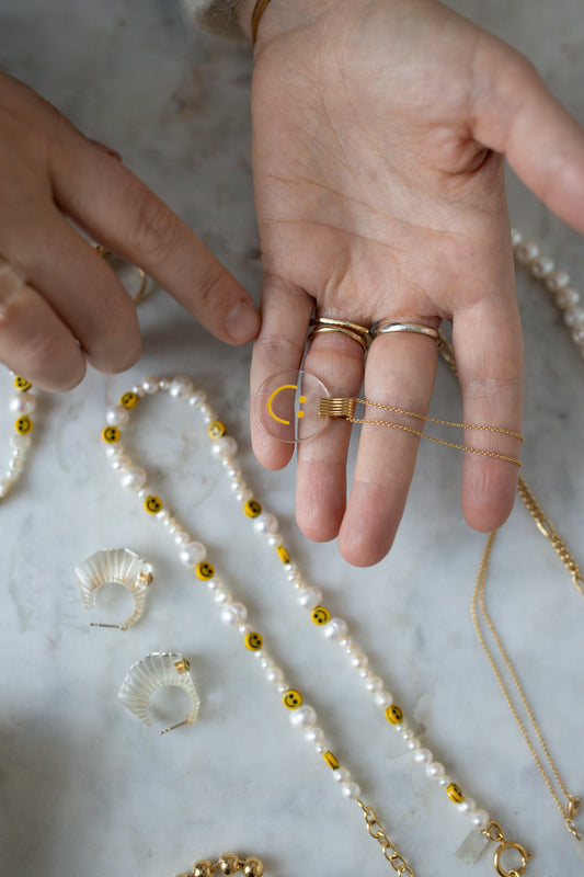 A hand is holding a WALD Berlin Smilie Dude Medaillon Gold Necklace, adorned with a gold crescent moon pendant. Various pieces of jewelry, including hand-painted polyester and pearl bead necklaces, are displayed on the marble surface.