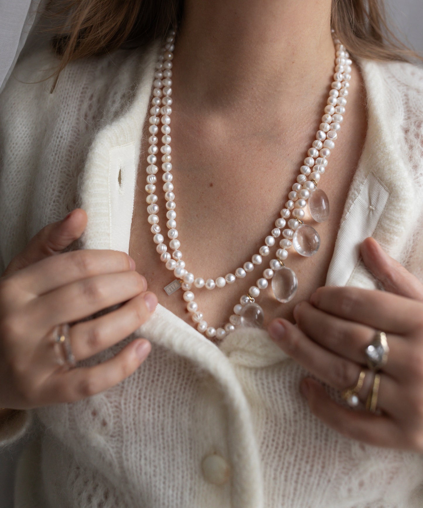 A woman in a cream cardigan layers her Happy Holidays Long Pearl Drop Necklace by WALD Berlin alongside berg crystal pendants, adjusting her necklaces with ring-adorned hands.