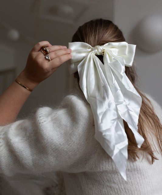 A person with long hair is preparing for the holidays by adjusting a WALD Berlin Happy Holidays Cream Hair Bow with one hand while wearing a cozy white sweater.