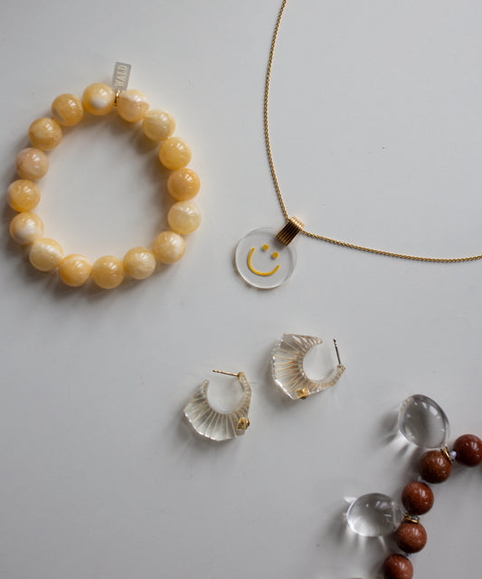 On a white surface, there's an 18k gold-plated Smilie Dude Medaillon Gold Necklace by WALD Berlin, complemented by a yellow beaded bracelet, clear hoop earrings, and a strand of mixed small beads.