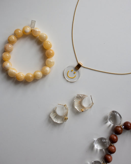 On a white surface, there's an 18k gold-plated Smilie Dude Medaillon Gold Necklace by WALD Berlin, complemented by a yellow beaded bracelet, clear hoop earrings, and a strand of mixed small beads.