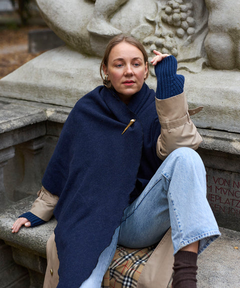 Eine Person, die einen „Homeland“-Schal von WALD Berlin x Helena Harfst trägt, an dem eine goldene Vogelbrosche befestigt ist, über einem beigen Trenchcoat und blauen Jeans, sitzt auf einem Steinsockel.